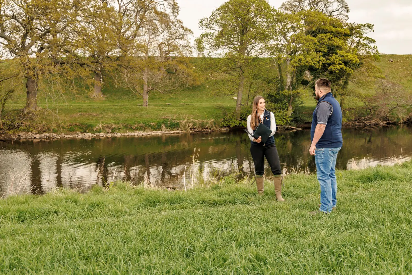 Farm consultant talking with client in grass field with river behind them